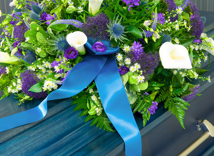 A colorful flower arrangement with white lilies, purple flowers, and a blue ribbon lies on top of a blue coffin situated in a formal setting.